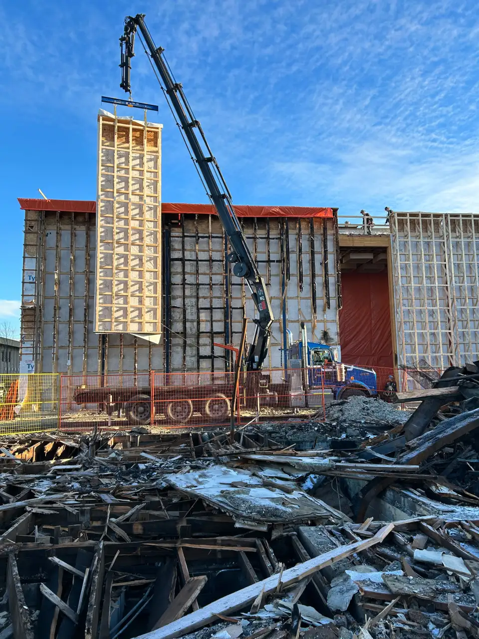 Lifting a new prefabricated wall panel into place. The damaged panels are behind, which were replaced one at a time. Note that the interior air/vapor membrane (Intello) in the damaged trusses is still intact &ndash; this is because of the mineral fiber insulation. The debris in the foreground is the building that exploded.